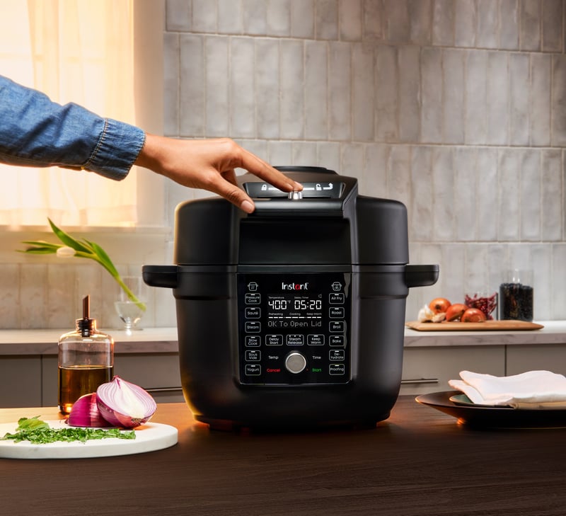 A hand reaches to press the control panel of a black Instant Pot multicooker on a kitchen counter