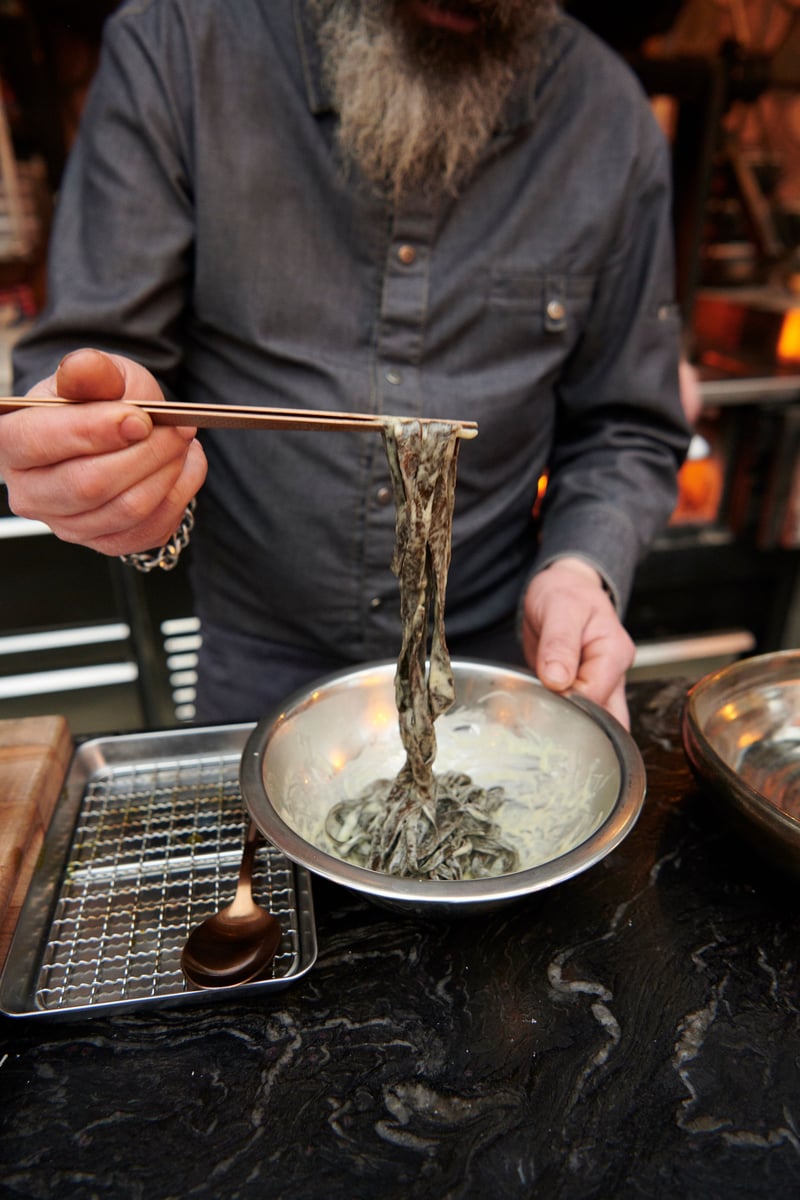 A bearded chef lifts noodles from a pan with chopsticks at a marble pass