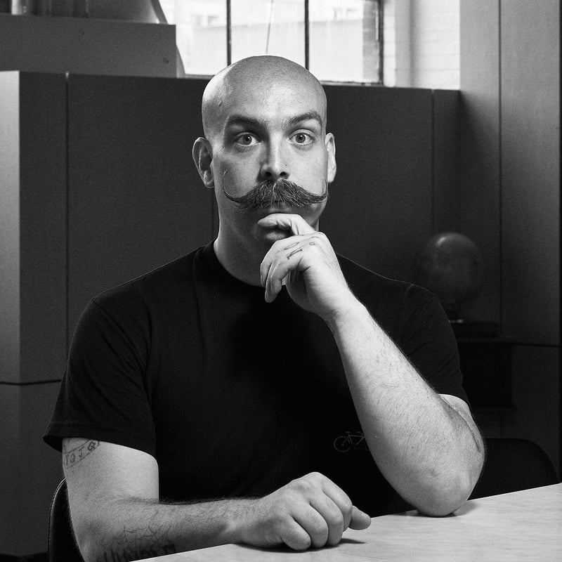A black and white portrait of a bald man with a heavy moustache, hand resting at his chin
