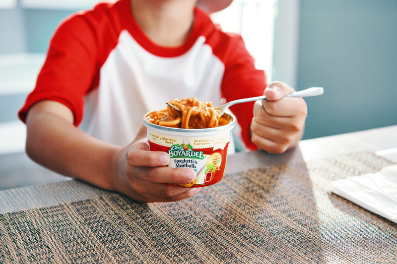 A child in a red baseball shirt eats pasta from a branded microwave cup at a table