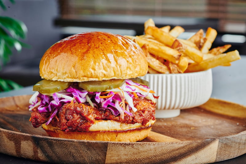 A fried chicken sandwich with slaw on a brioche bun, served on a board with a bowl of fries