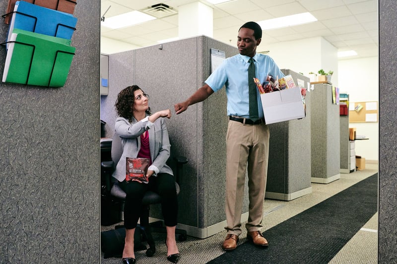 Two colleagues meet in an office cubicle aisle, one handing across a carton of food