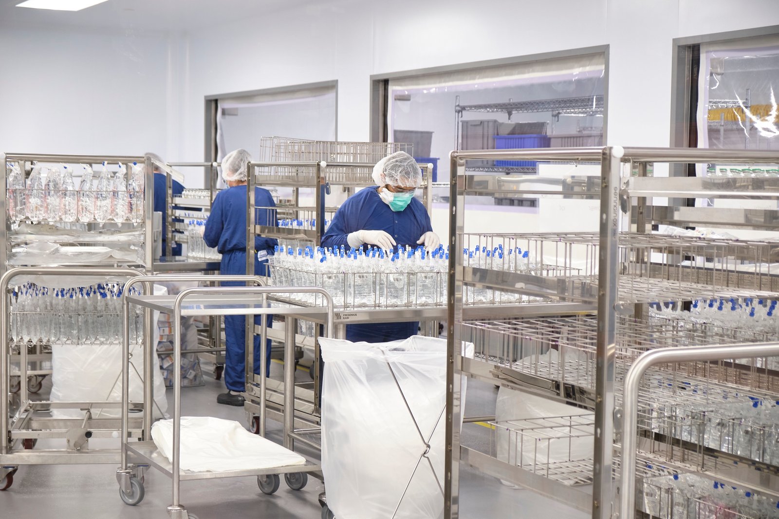 Workers in blue scrubs and hairnets moving steel racks through a clean pharmaceutical production room