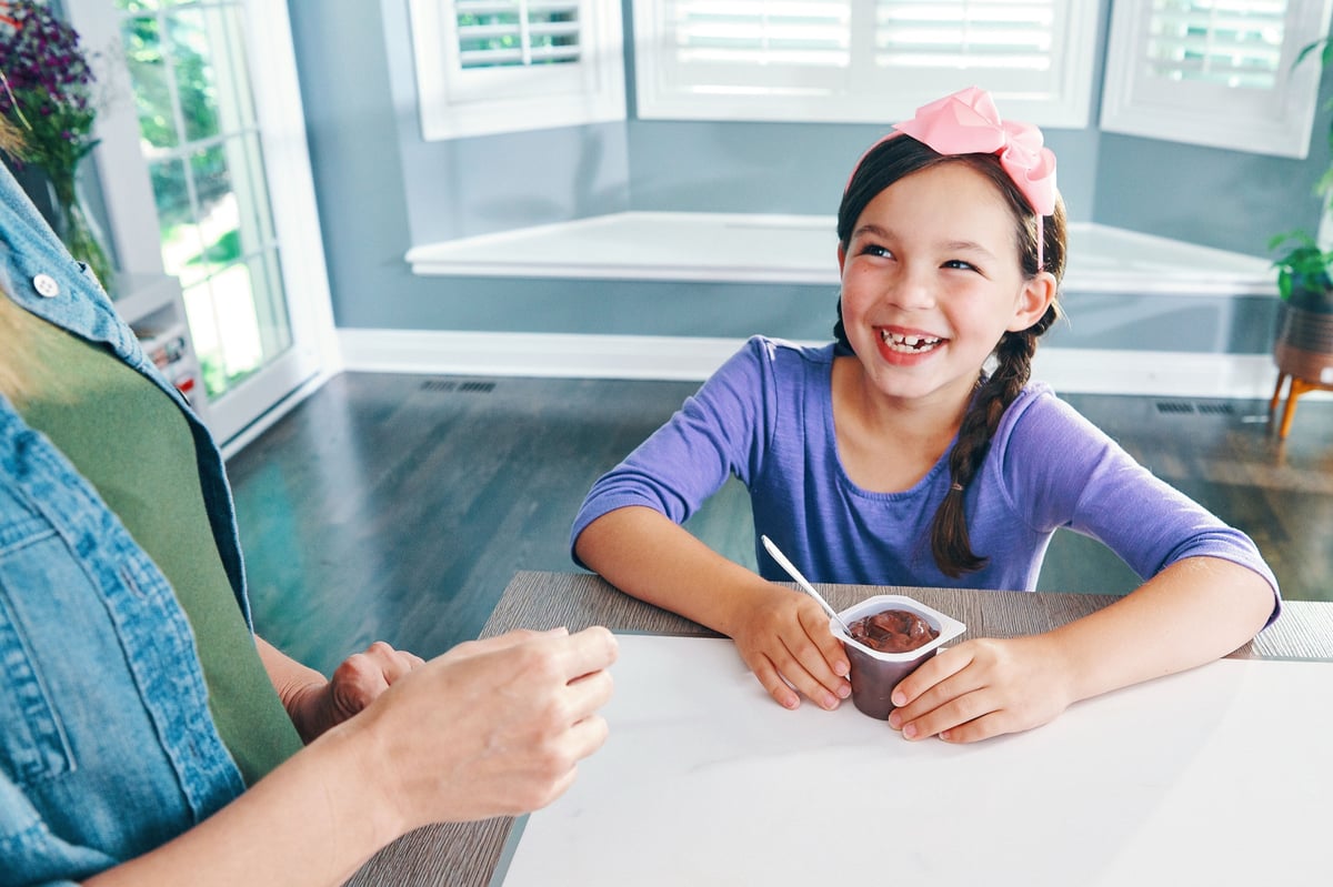 A young girl in a pink headband smiles across a kitchen table, a small cup in her hands and an adult's hands opposite her