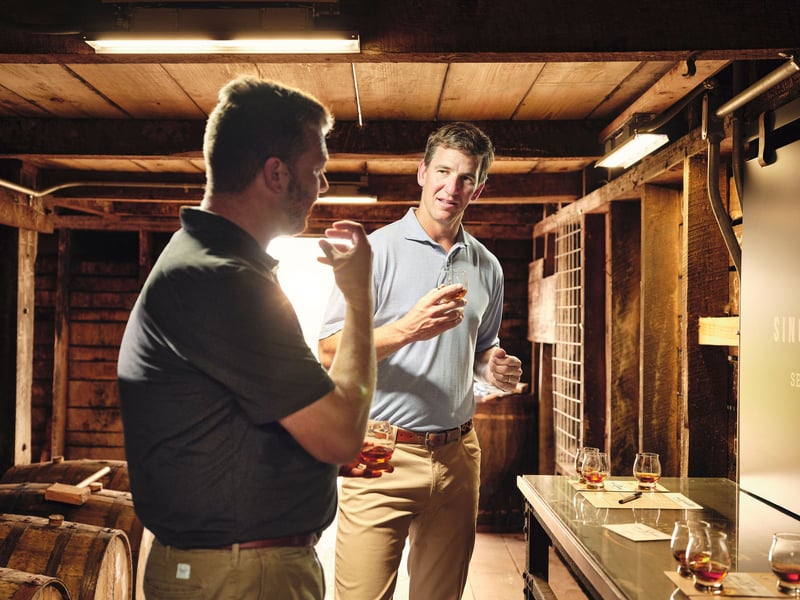 Two men stand talking in a timber rickhouse, warm strip lights overhead