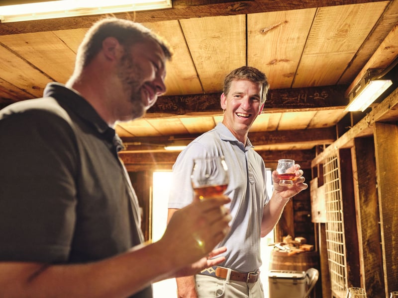 Two men stand talking in a rickhouse, each holding a glass of bourbon