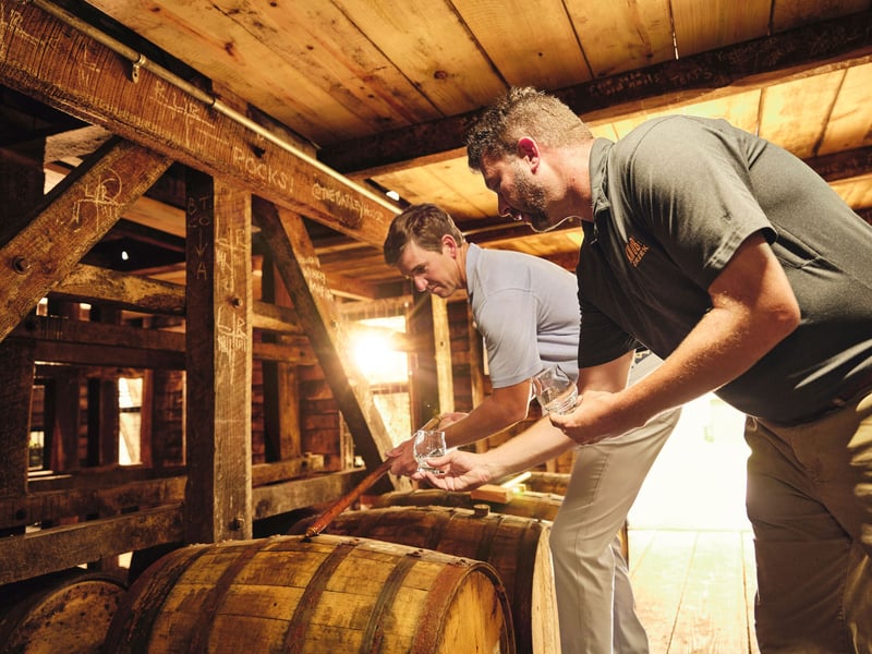 Two men in a timber rickhouse lean over a bourbon barrel, warm light coming through behind them