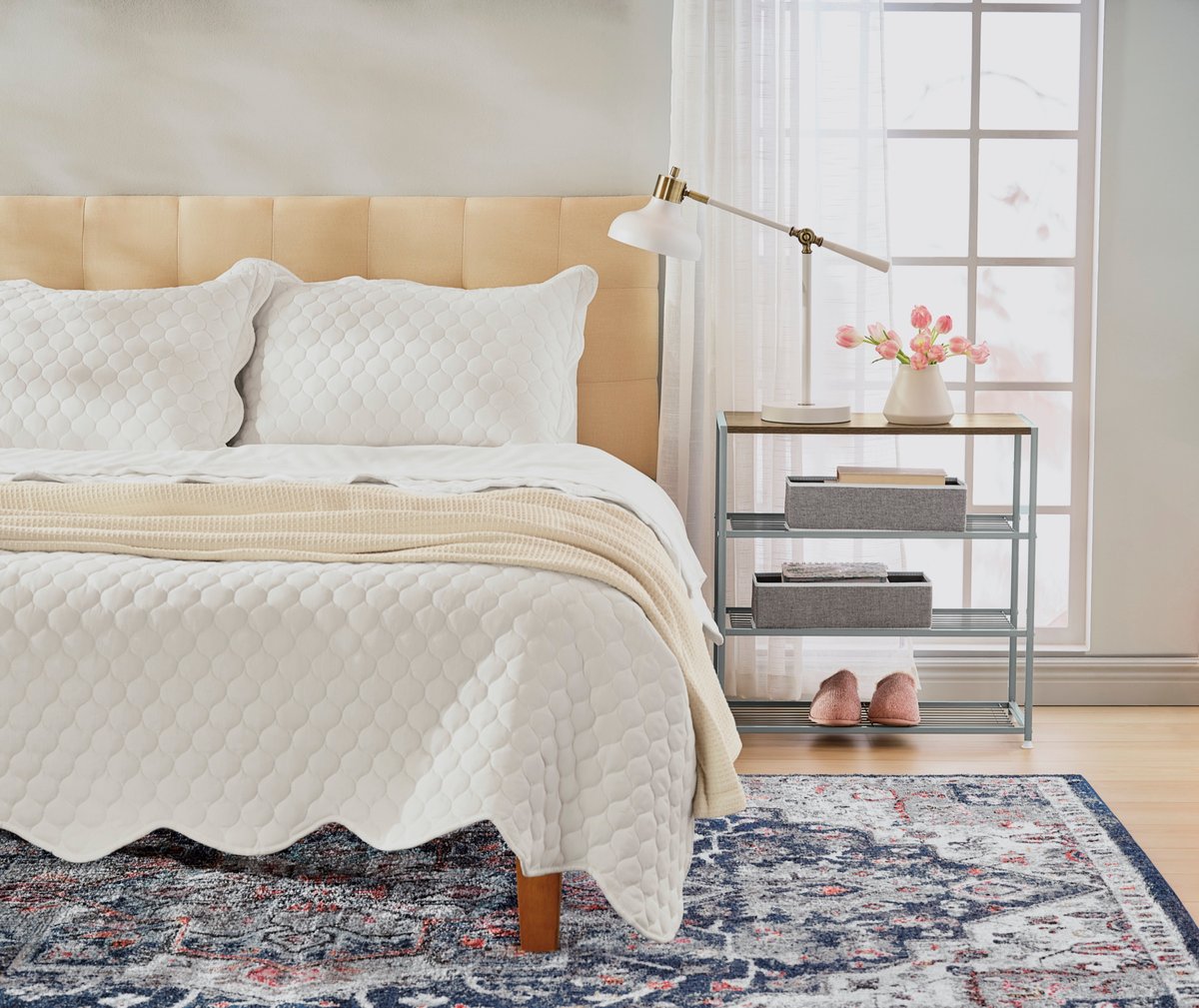 A cream bedroom with a quilted bed, patterned rug and slippers by a bright window