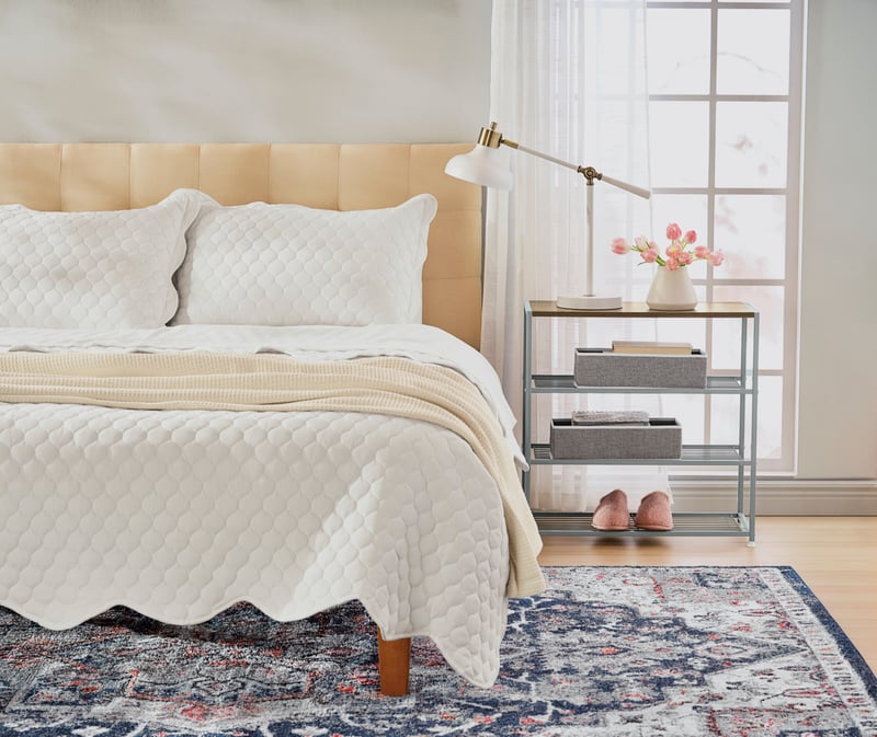 A cream bedroom with a quilted bed, patterned rug and slippers by a bright window