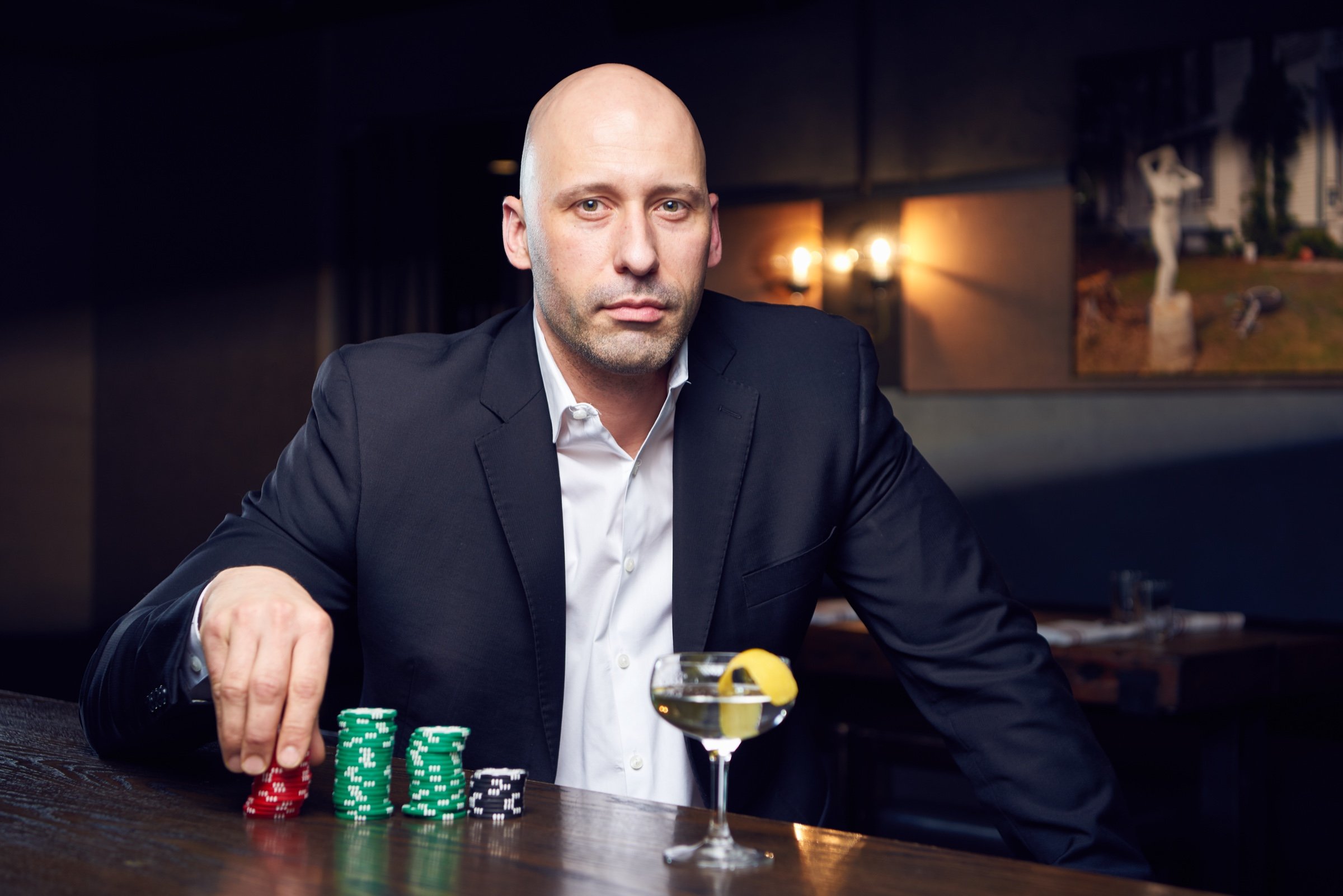 A man in a dark suit sits at a bar with poker chips stacked in front of him and a cocktail at his elbow