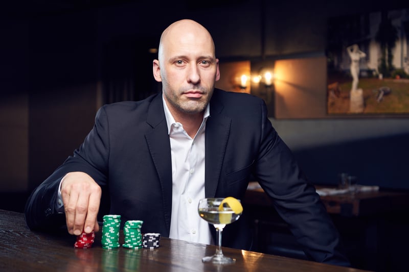 A man in a dark suit sits at a bar with poker chips stacked in front of him and a cocktail at his elbow