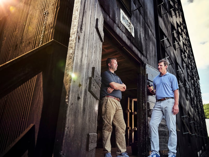 Two men stand outside a dark timber distillery building, hands at their sides