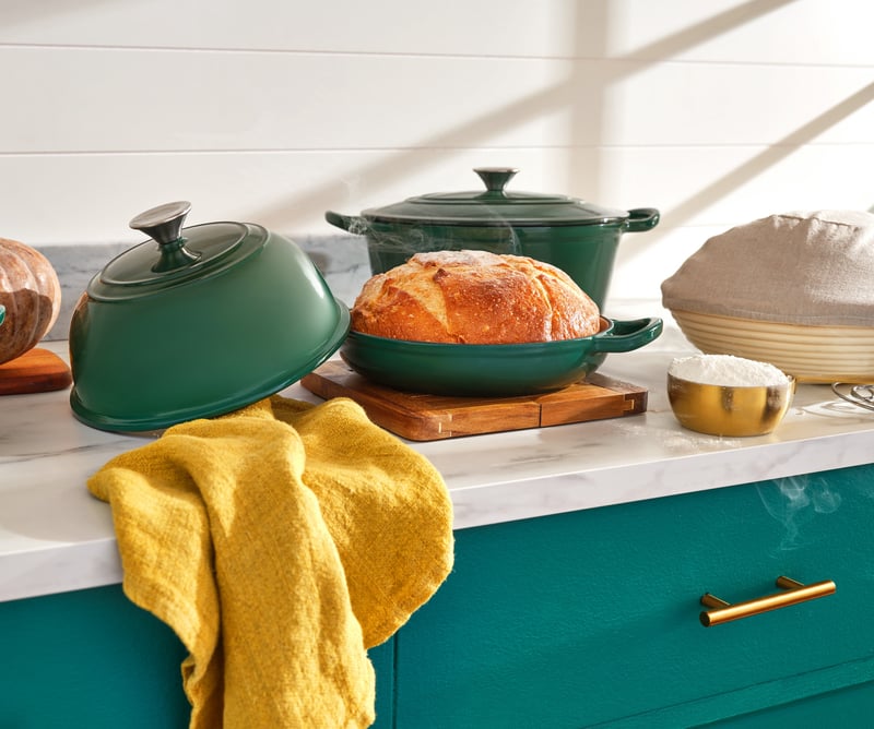 Green enamel casserole dishes and a fresh loaf on a teal kitchen island with a yellow cloth