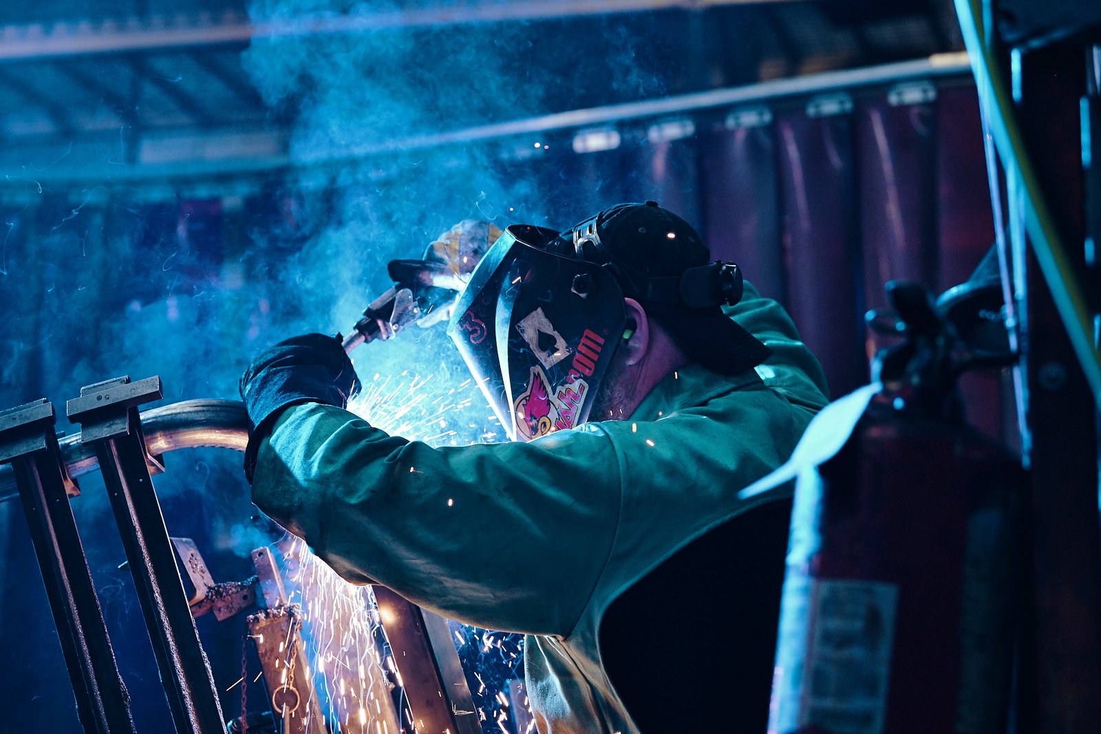 A welder in a dark workshop, sparks flaring from the torch in a haze of blue smoke