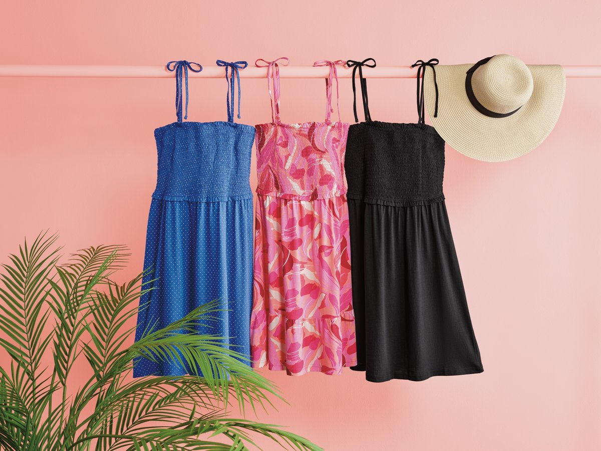 Summer dresses and a straw hat hanging on a rail against a pink wall, palm fronds in the corner