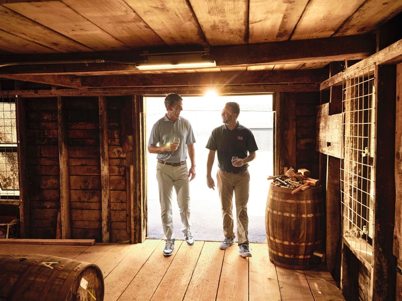 Two men walk through a rickhouse toward the camera, daylight flooding the doorway behind them
