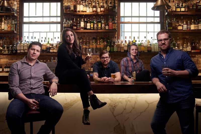 Five people gathered along a bar counter, tall windows and backlit bottles behind them