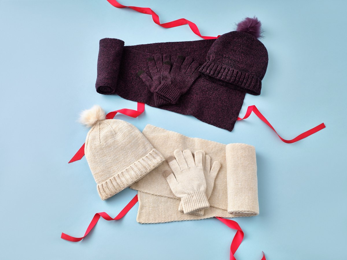 A knitted bobble hat, scarf and gloves arranged on a pale blue ground with red ribbon
