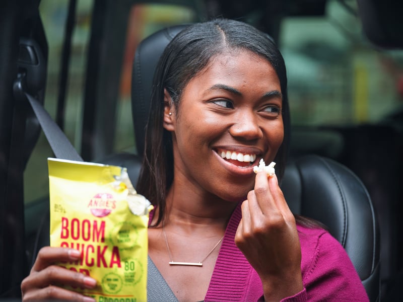 A woman in a car seat laughs while eating popcorn from a bright yellow bag
