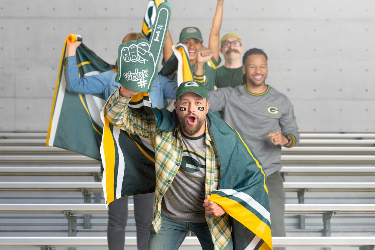 Fans in green and gold cheer on stadium bleachers, one holding a foam finger