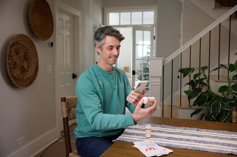 A man sits at a dining table in a bright hallway, phone in hand and paperwork spread in front of him