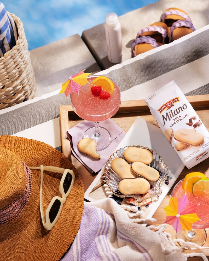 A picnic tray with a pink cocktail, Milano biscuits and sunglasses on a striped blanket