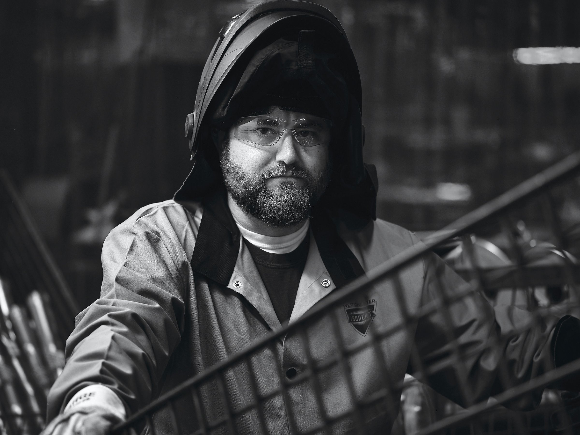 A black and white portrait of a bearded worker in a hood, lit hard against a dark industrial background