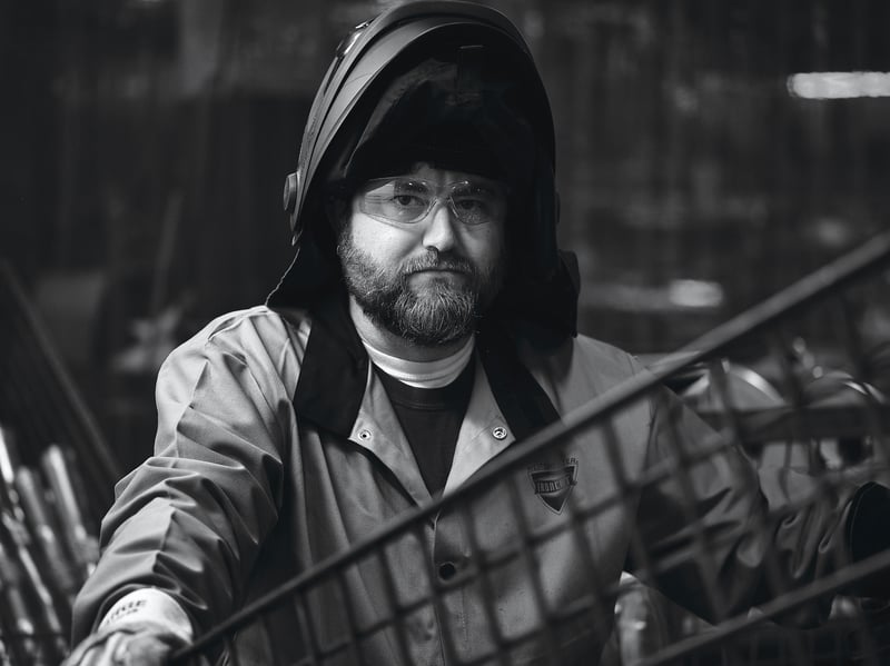 A black and white portrait of a bearded worker in a hood, lit hard against a dark industrial background