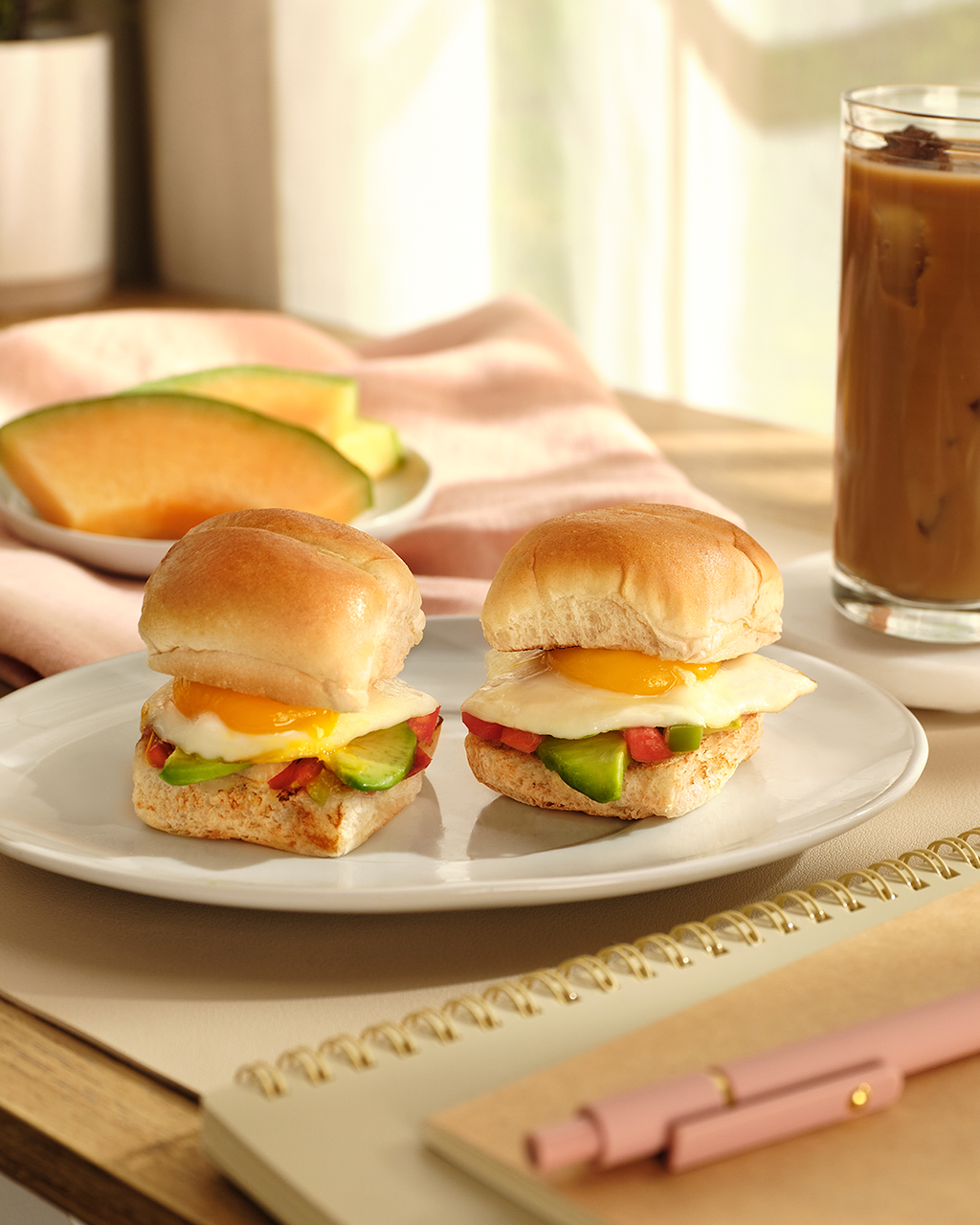 Two breakfast sandwiches on soft rolls on a white plate, iced coffee, melon and a notebook in warm window light