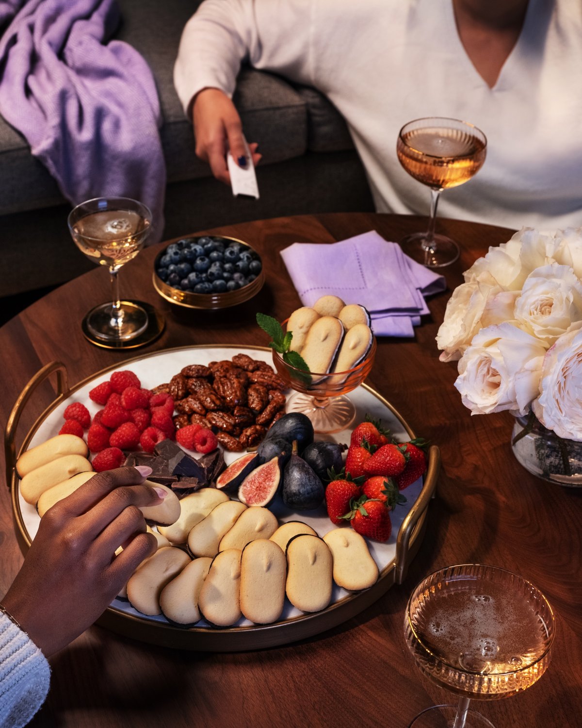 Two people reach across a dark wooden table laid with a grazing board of biscuits, figs, berries and glasses of wine