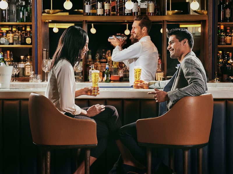 Three people talking and drinking at a warmly lit bar, backlit bottles on the shelves behind them