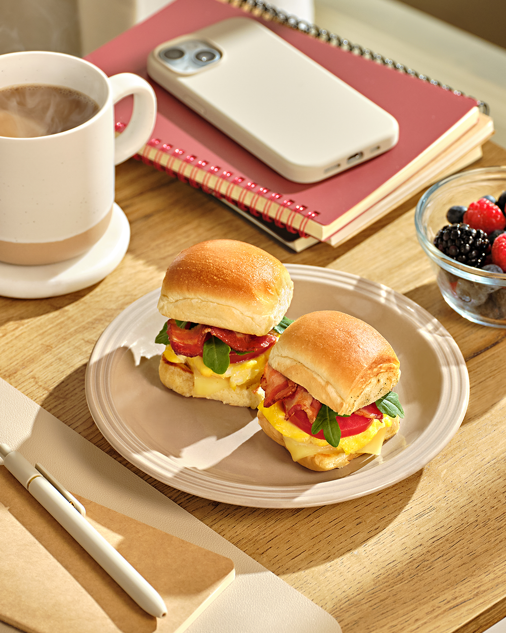 Two breakfast sliders on a plate beside a coffee cup, berries, a notebook and a phone on a wooden desk