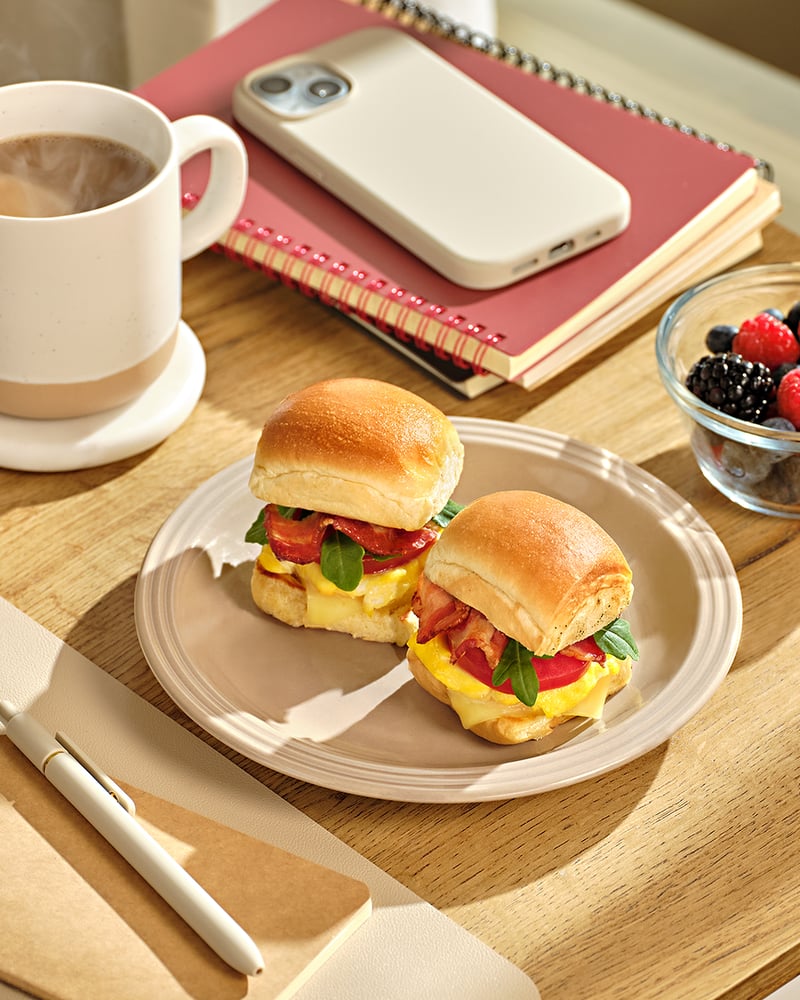 Two breakfast sliders on a plate beside a coffee cup, berries, a notebook and a phone on a wooden desk