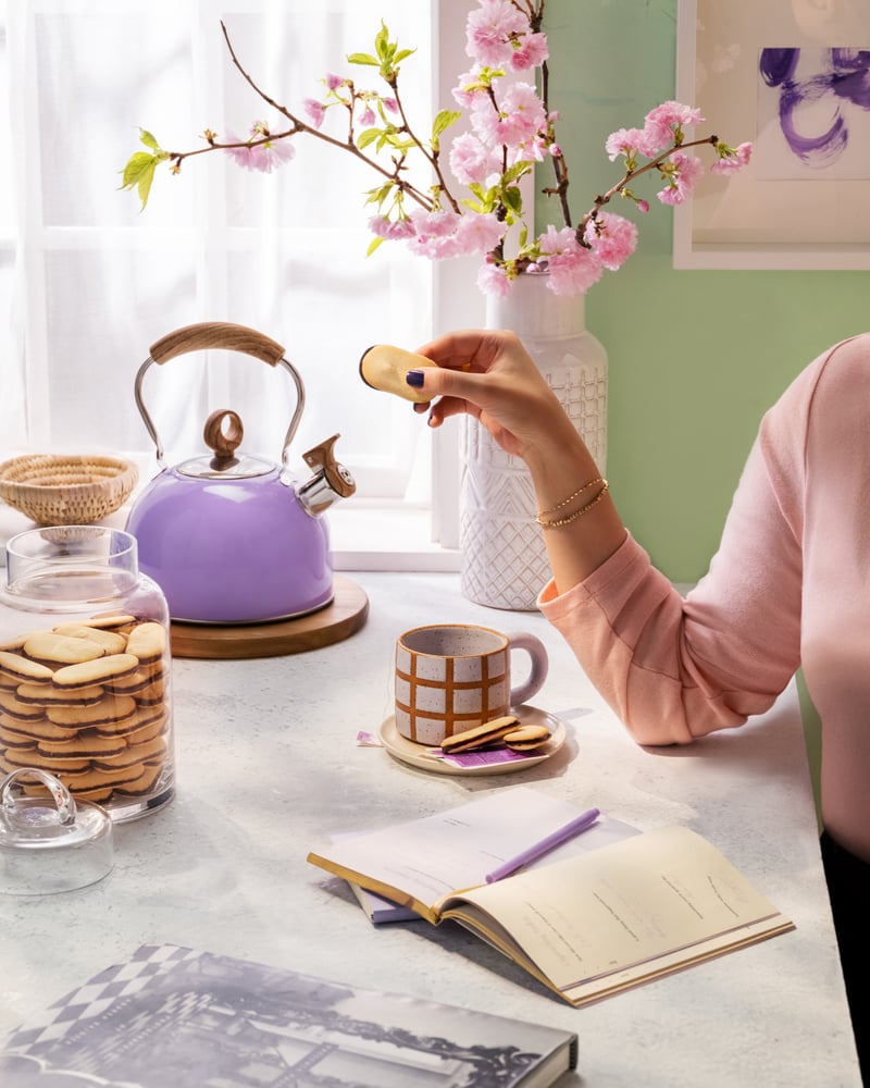 A hand reaches for a biscuit at a table set with a lilac kettle, teacups and an open notebook, cherry blossom behind