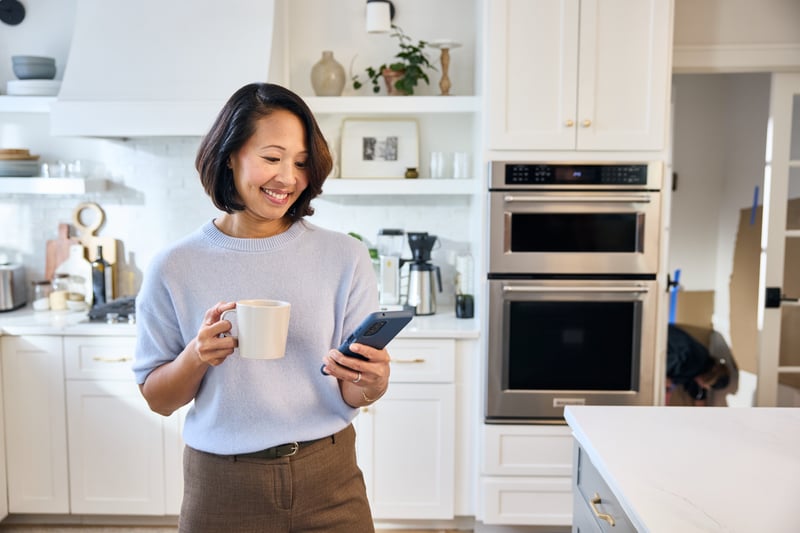 A woman stands in a bright white kitchen holding a mug in one hand and her phone in the other