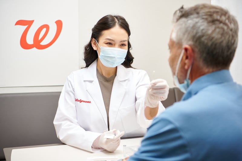 A pharmacist in a white coat and mask speaks with an older customer across a Walgreens counter
