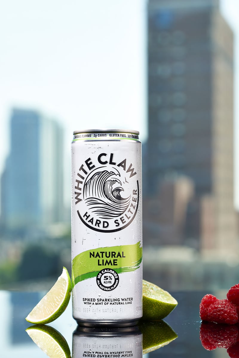 A can of White Claw Natural Lime stands on a reflective surface with lime wedges, a city skyline behind