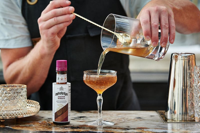 A bartender strains a cocktail into a coupe glass at a marble bar, the bitters bottle standing beside the shaker