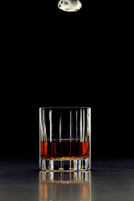 A cut-glass tumbler of whisky on a dark reflective bar as a large ice cube drops toward it