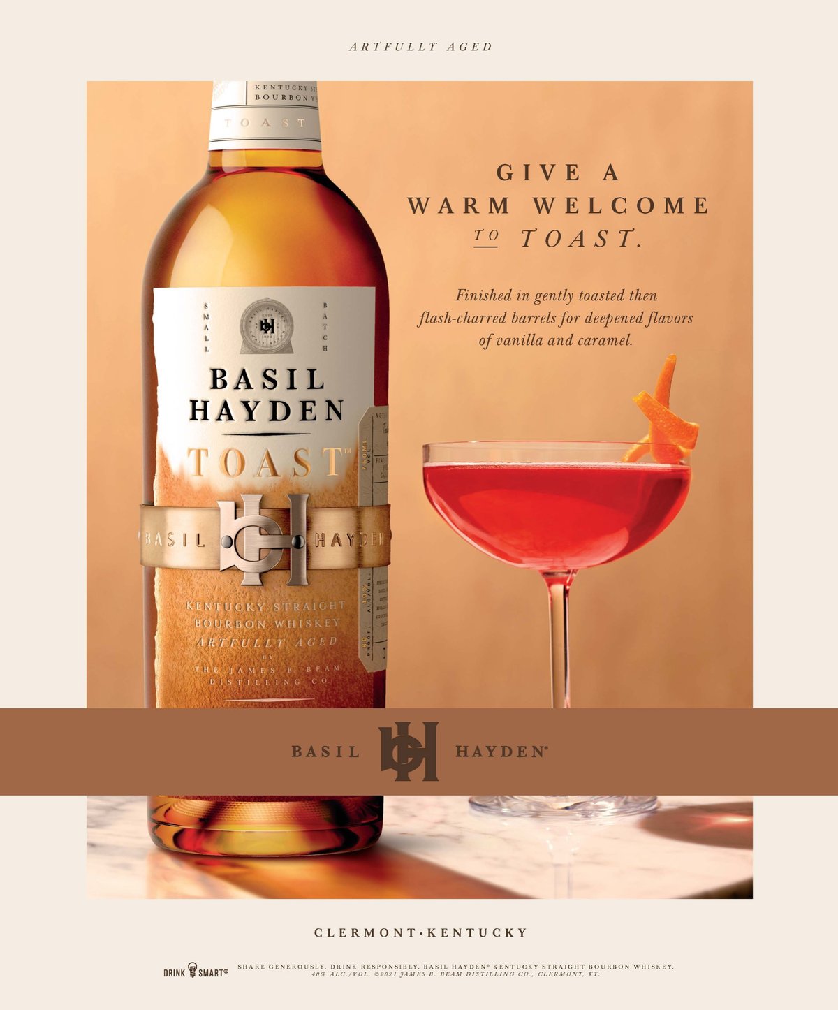 A Basil Hayden Toast bottle beside a red cocktail in a coupe on a warm sand-colored layout