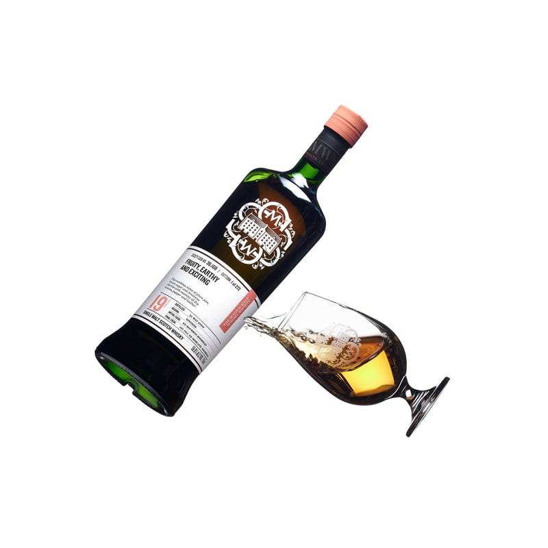 A whisky bottle lying on its side pouring into a small tasting glass, photographed on white