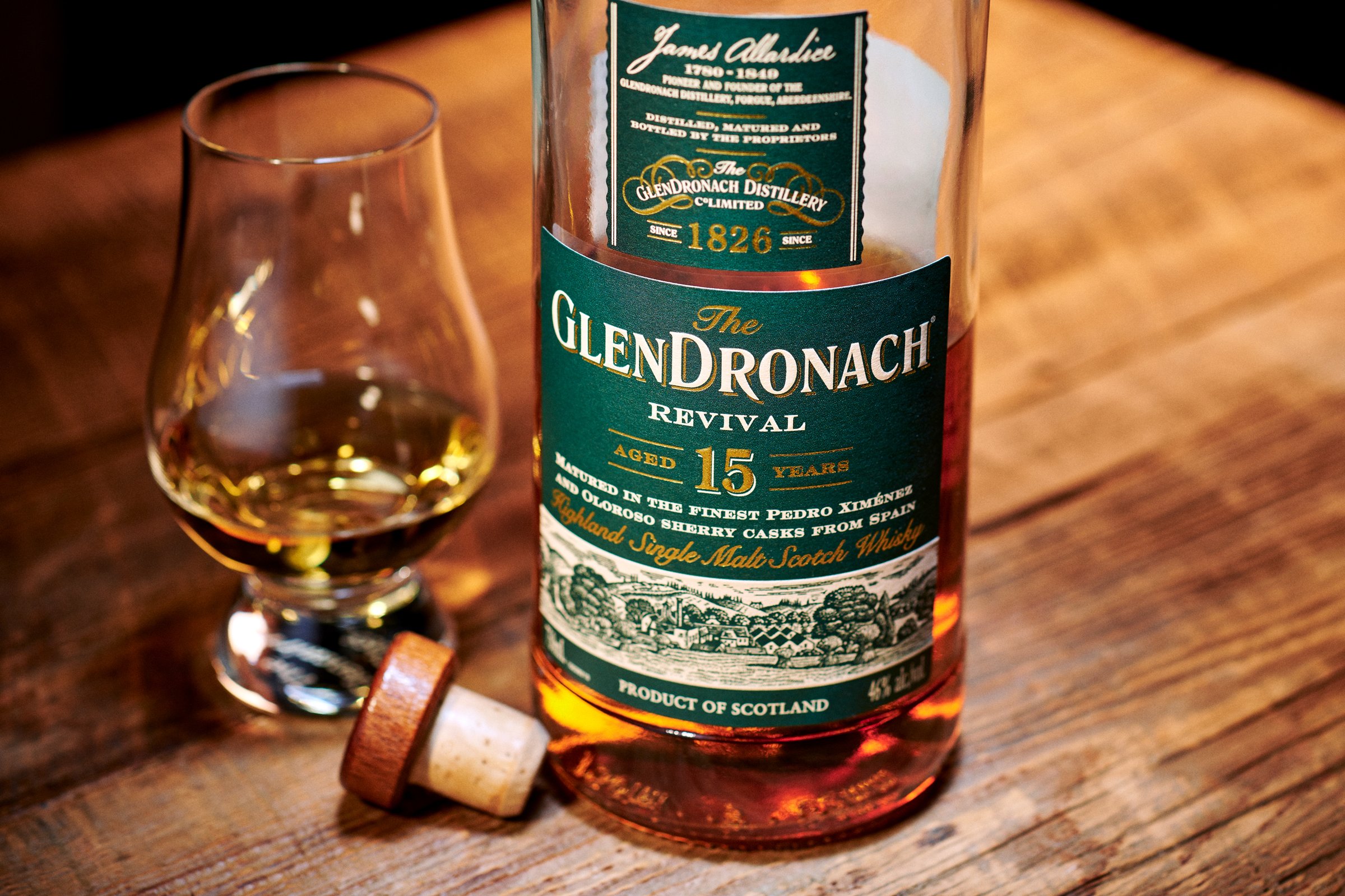 A bottle of GlenDronach 15 beside a Glencairn glass of whisky on a wooden tabletop, cork resting alongside