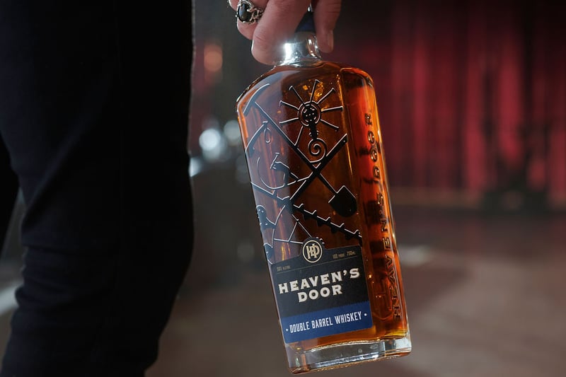A hand lifts a decorated Heaven's Door whiskey bottle from a dark surface under red light