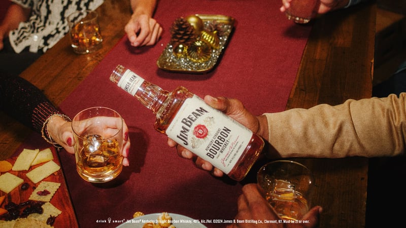 Hands reaching across a red table for glasses of bourbon, one holding a Jim Beam bottle