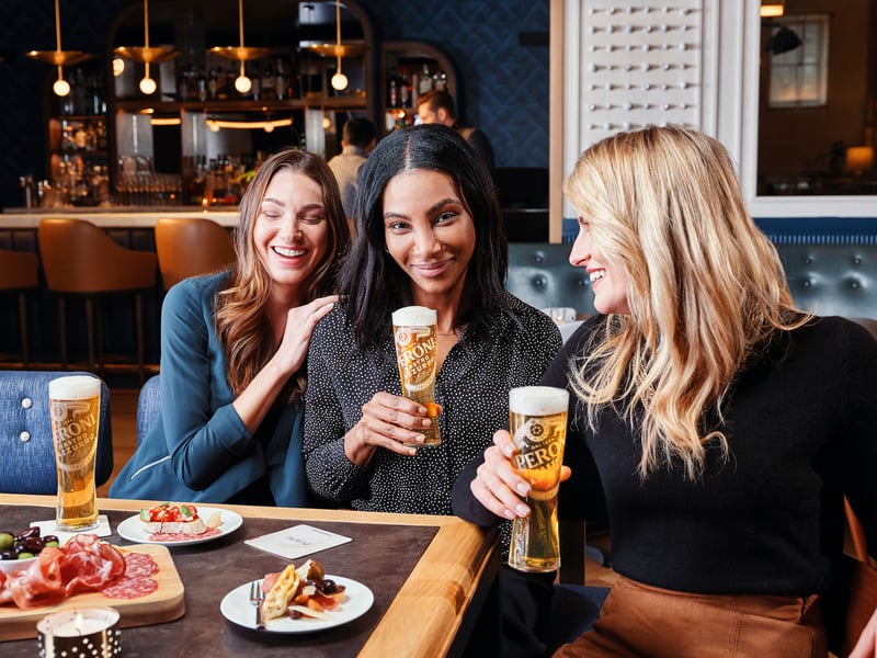 Three women laugh together at a bar with tall glasses of lager and a plate of food in front of them