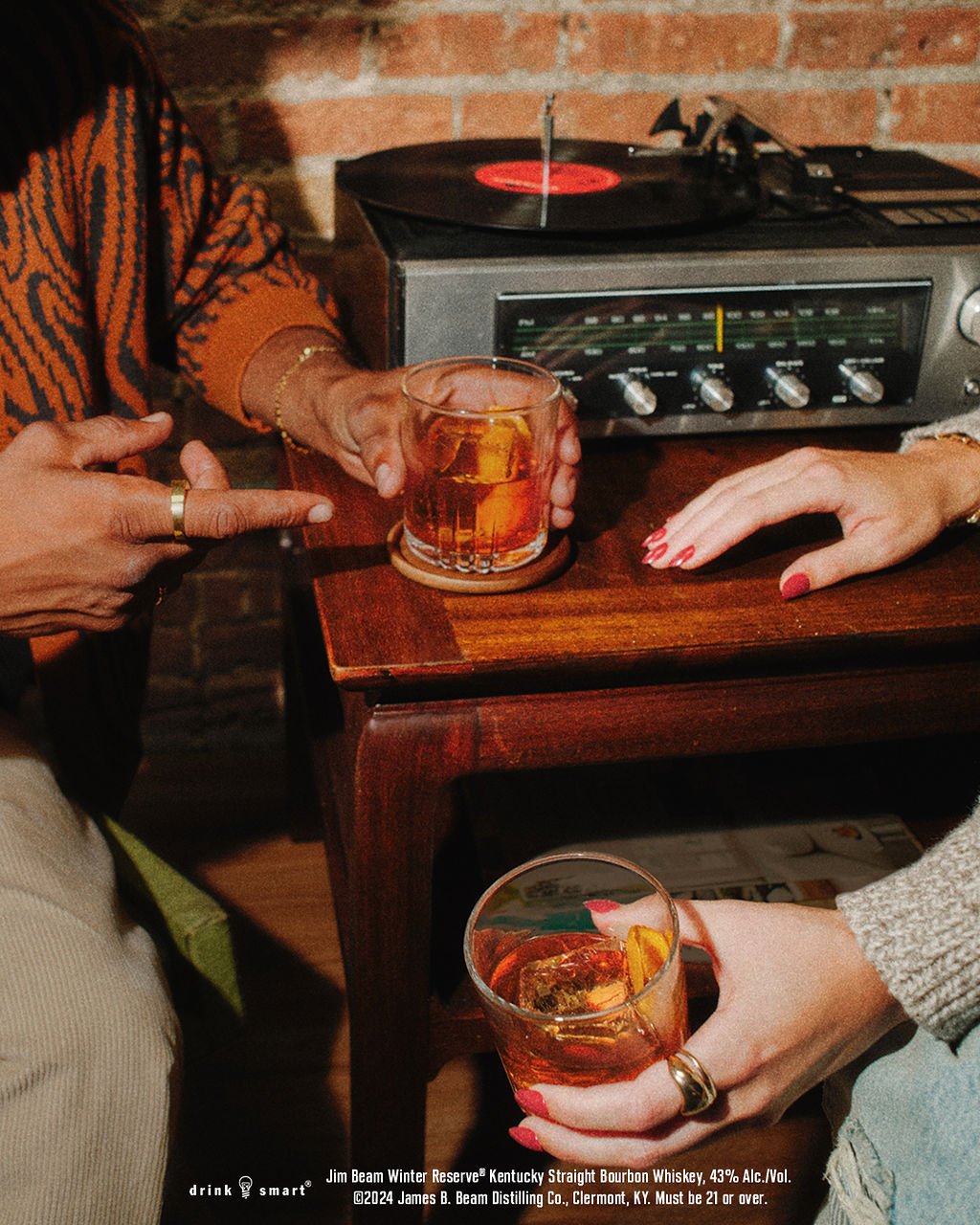 Hands lift glasses of bourbon from a wooden side table beside a record player