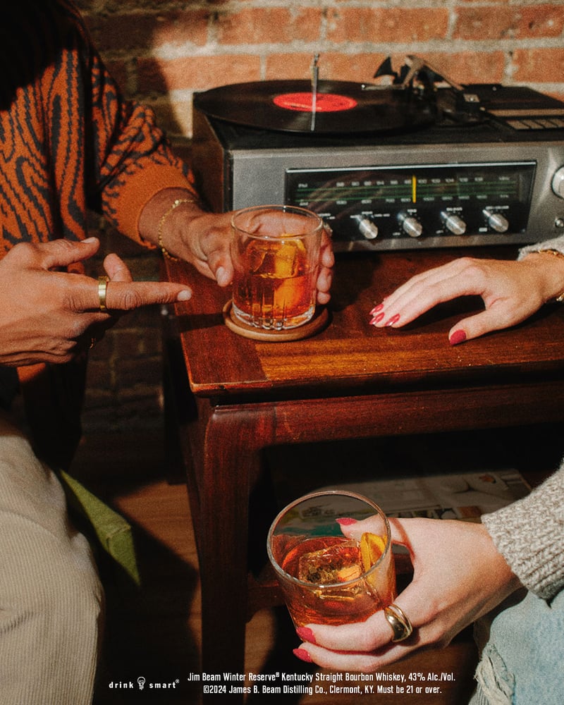 Hands lift glasses of bourbon from a wooden side table beside a record player