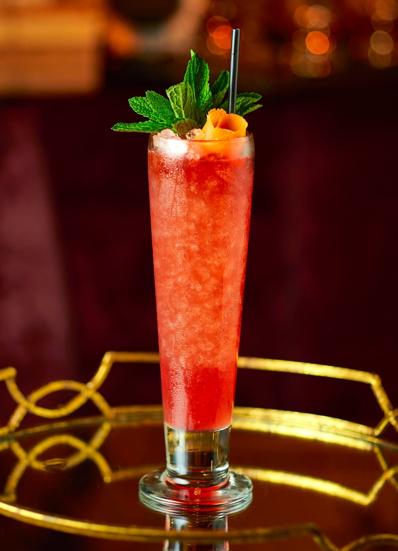 A tall red cocktail with mint and citrus on a gold tray against a deep red backdrop