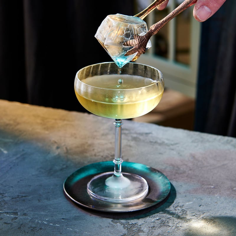 A hand lowers a large clear ice cube into a coupe of pale yellow cocktail with tongs