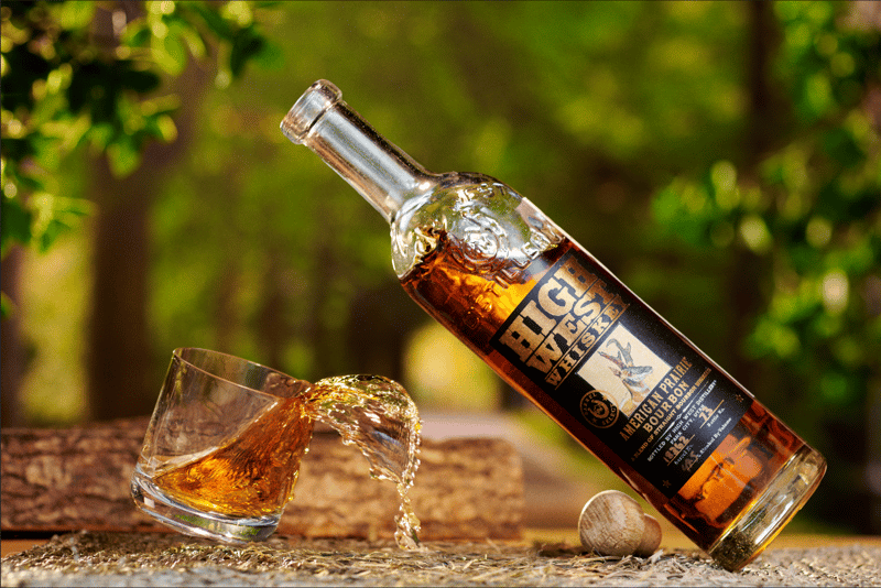 A whisky bottle tipped mid-pour into a glass on a stone ledge, dappled green foliage behind
